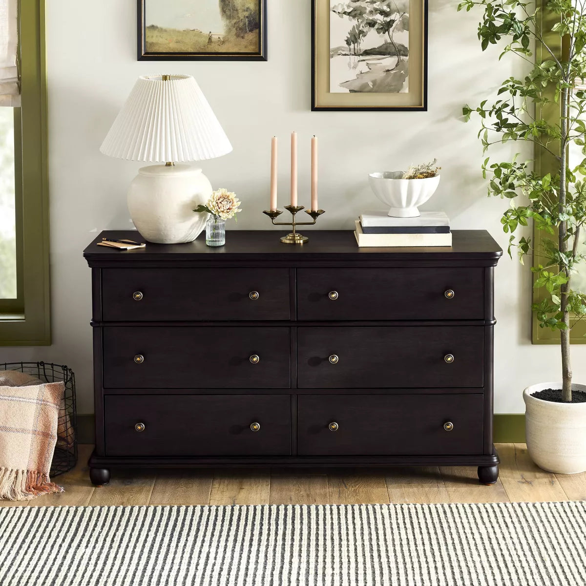 A dark brown colored dresser with six drawers, featuring round knobs, is adorned with a white lamp, books, a bowl, and a candle holder on top Black.