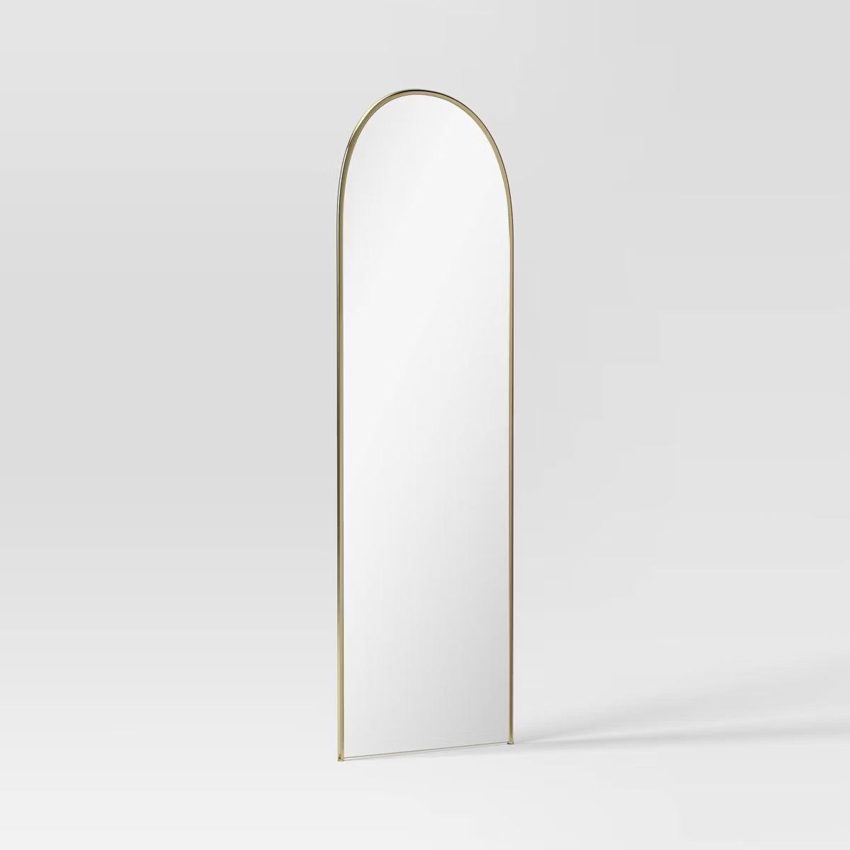 An arched mirror with a thin gold colored frame against a light gray background.