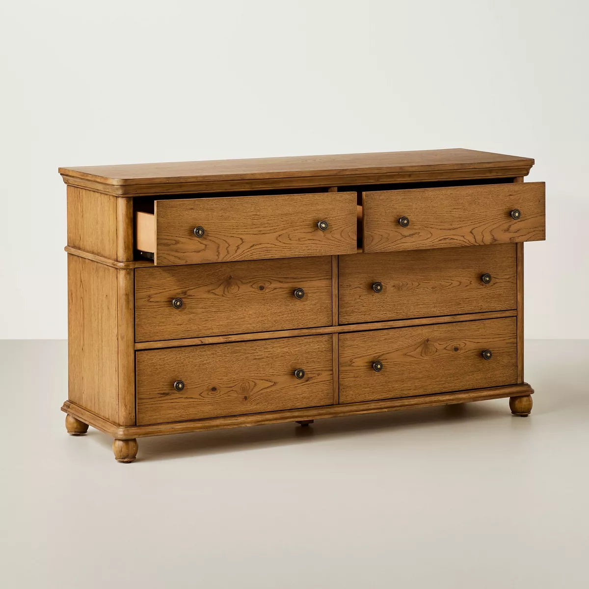 A dark brown colored dresser with six drawers, featuring round knobs, is adorned with a white lamp, books, a bowl, and a candle holder on top Aged Oak.