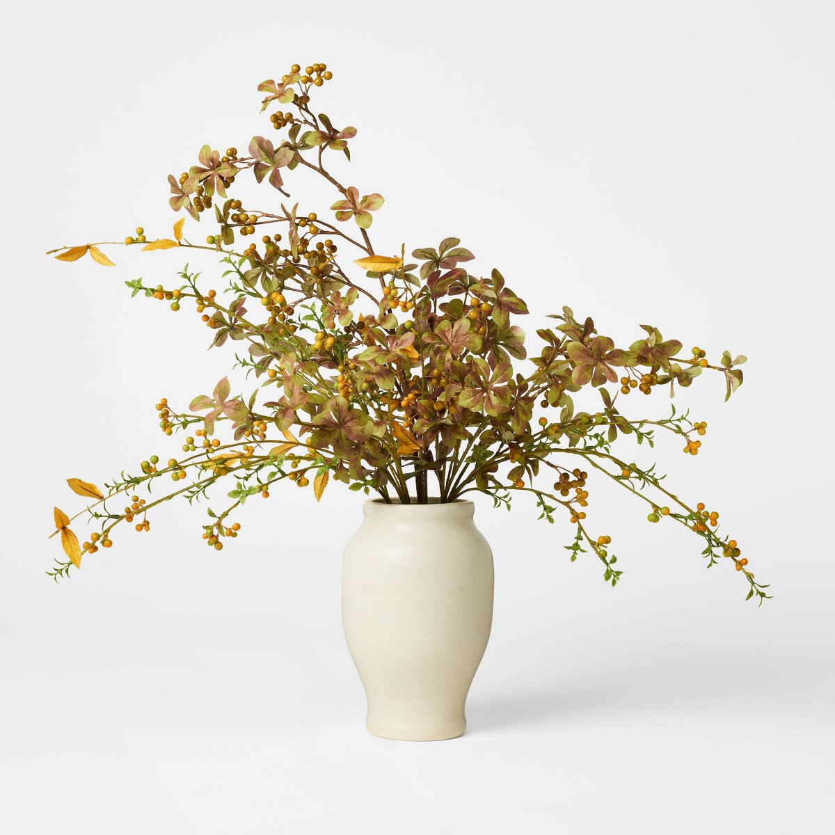An artificial autumn leafy branch arrangement in a matte cream ceramic vase, photographed on a clean white studio background.