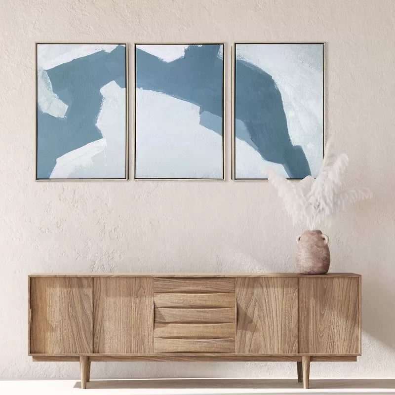 Three abstract blue artworks on a wall above a wooden cabinet.