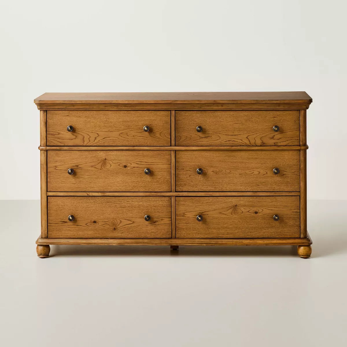 A dark brown colored dresser with six drawers, featuring round knobs, is adorned with a white lamp, books, a bowl, and a candle holder on top Aged Oak.