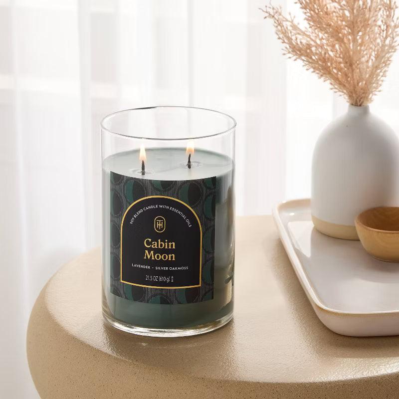Candle labeled 'Cabin Moon' on a table with a vase and decorative items in the background 21.5oz