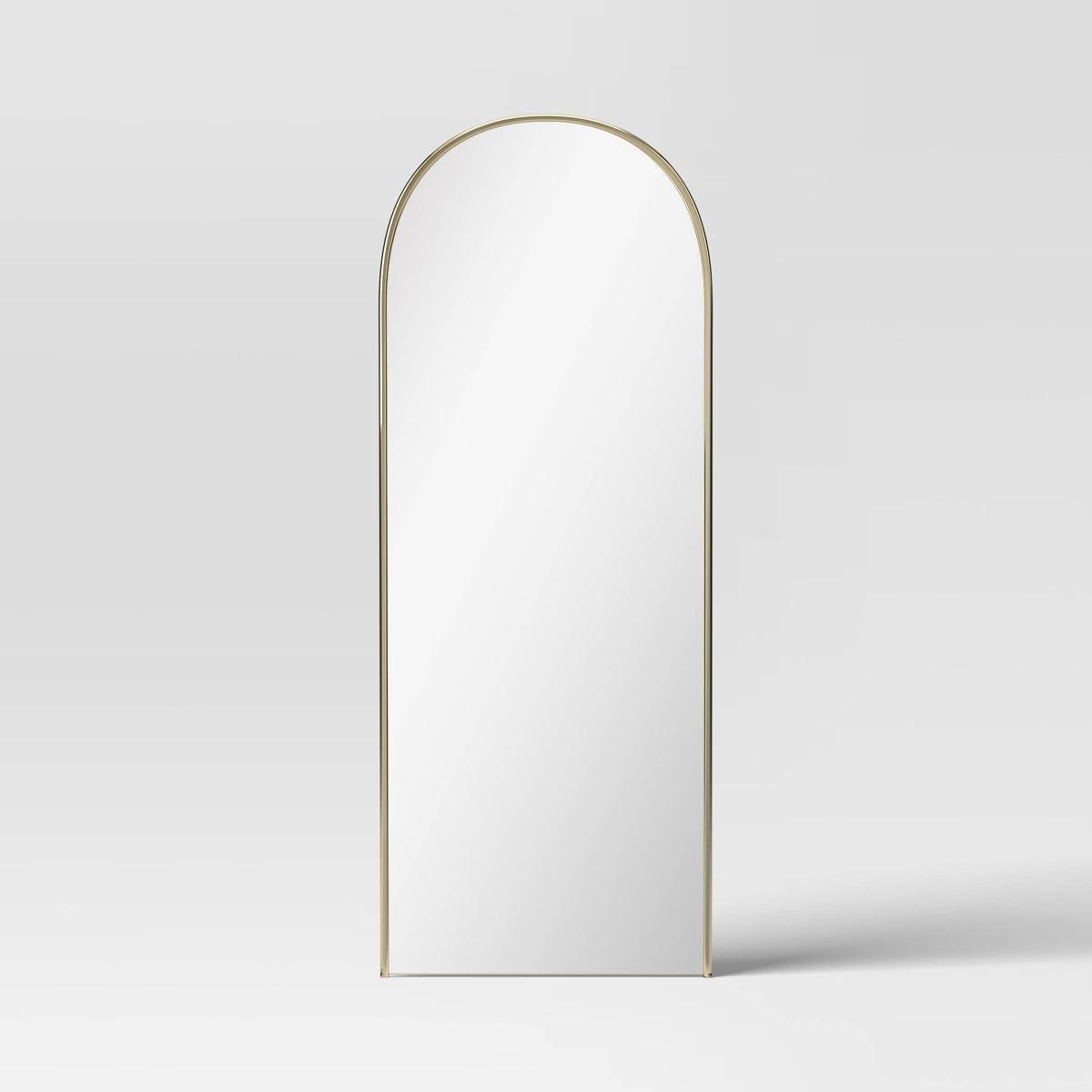An arched mirror with a thin gold colored frame against a light gray background.