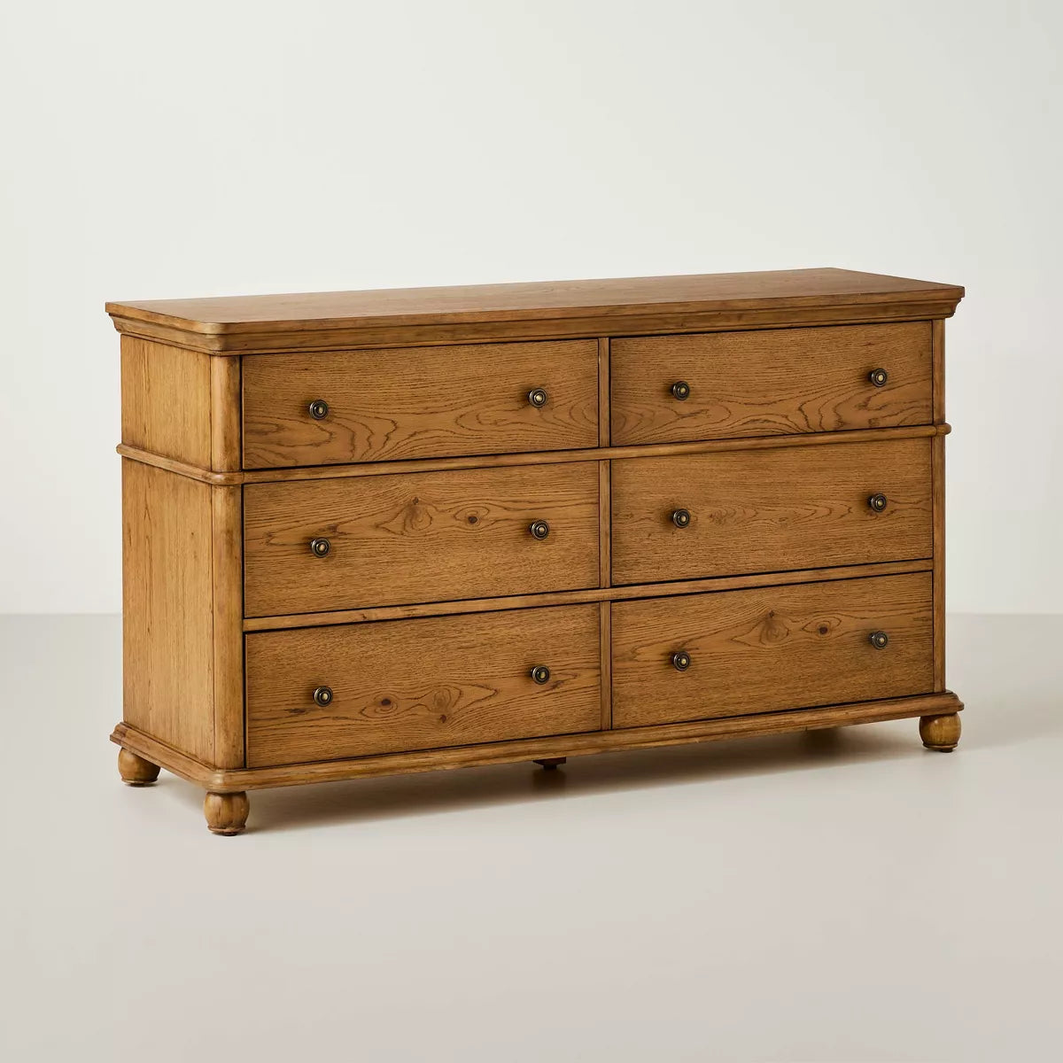 A dark brown colored dresser with six drawers, featuring round knobs, is adorned with a white lamp, books, a bowl, and a candle holder on top Aged Oak.