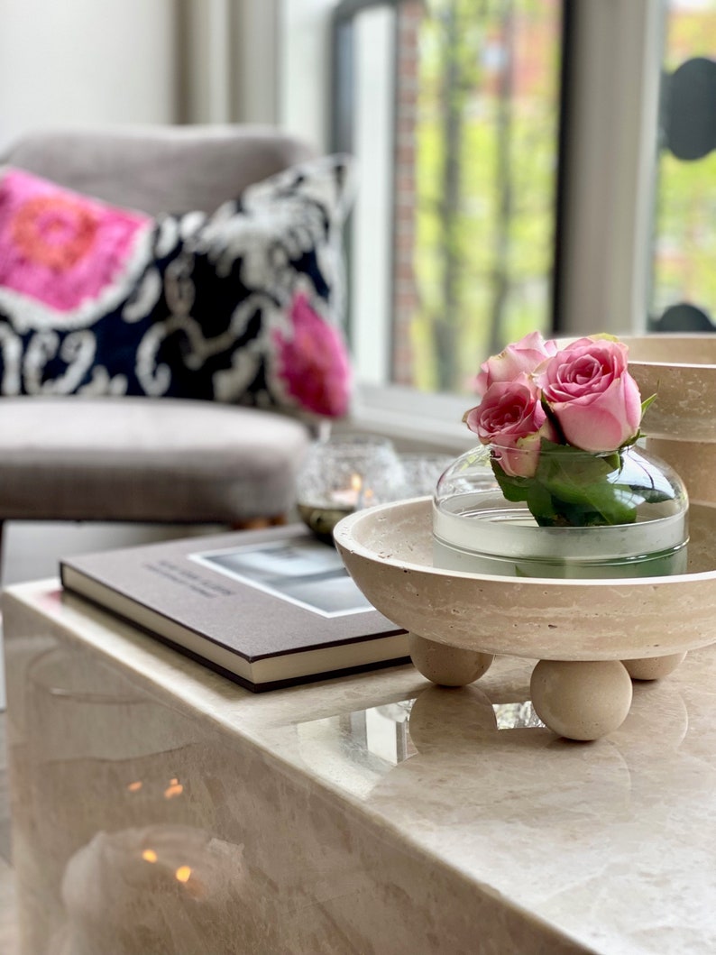 Decorative setting with a vase of pink roses on a marble table, surrounded by books and a cushioned chair.
