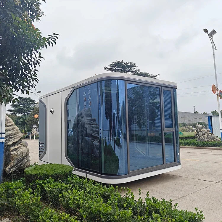 A modern small modular office or guardhouse with a white exterior and large dark-tinted glass windows, photographed outdoors on a paved area with shrubs, trees, and a cloudy sky in the background.