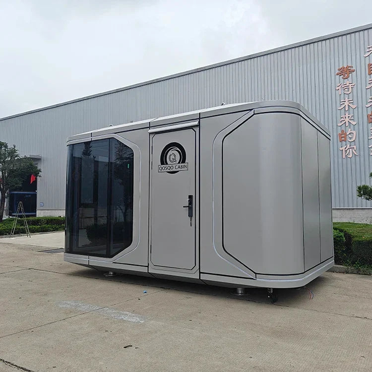 A modern modular outdoor toilet container with a sleek silver-gray exterior, dark tinted glass panels, and a door featuring a circular logo, parked outdoors in front of a large corrugated metal warehouse building.