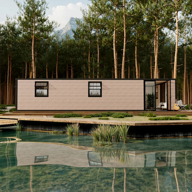 A modern single-story prefab cabin with a light beige brick exterior and black trim, situated on a wooden deck beside a reflective pool, with a dense pine forest and mountains in the background.