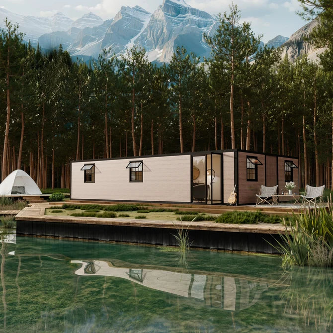 A modern rectangular mobile home with a light beige exterior and black trim sits on a wooden dock beside clear water, surrounded by tall evergreen trees and snow-capped mountains in the background.