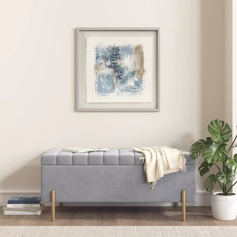 Gray upholstered bench with a blanket in a room with a plant and abstract art on the wall.