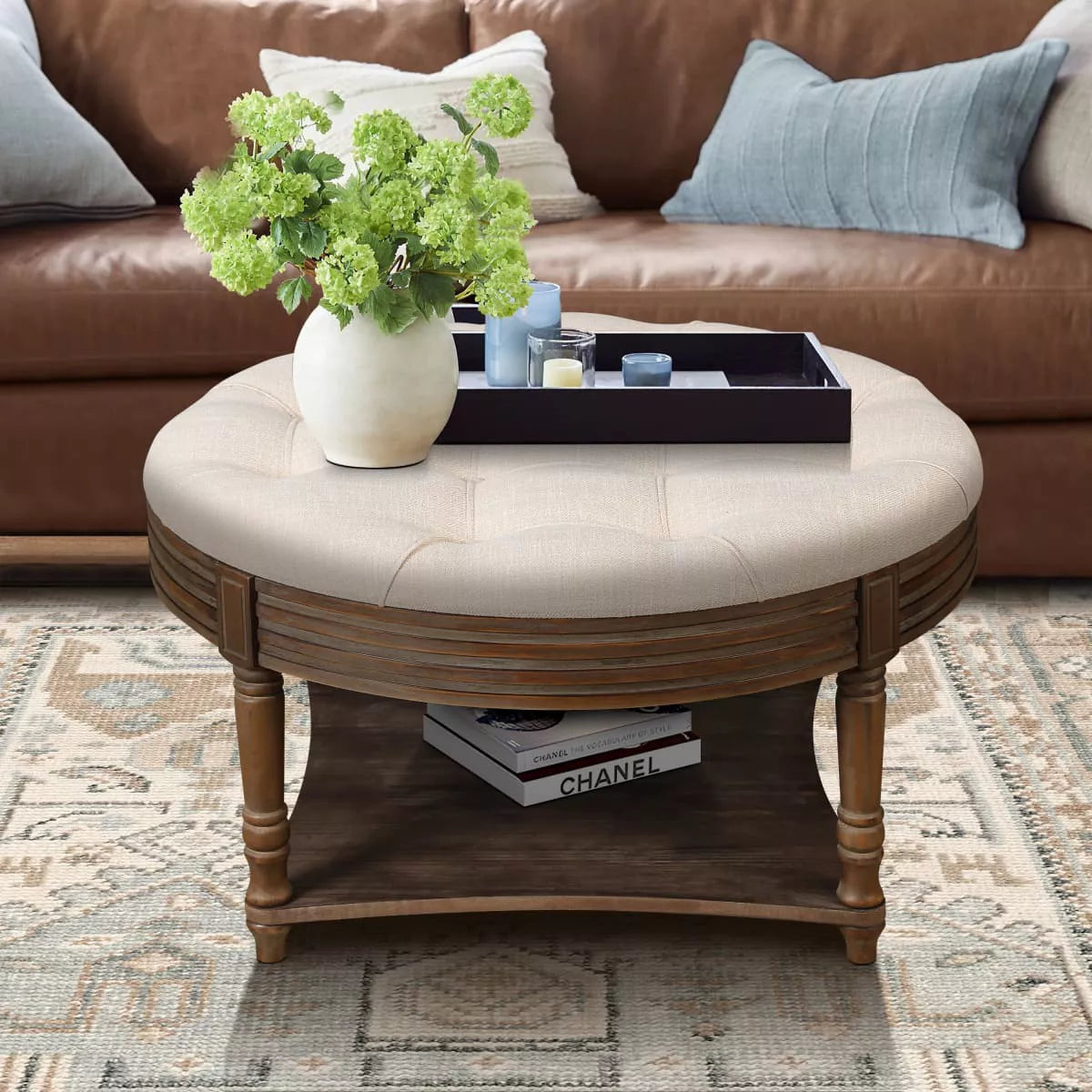 Round wooden coffee table with a beige cushion, vase of green flowers, and books on a patterned rug. beige