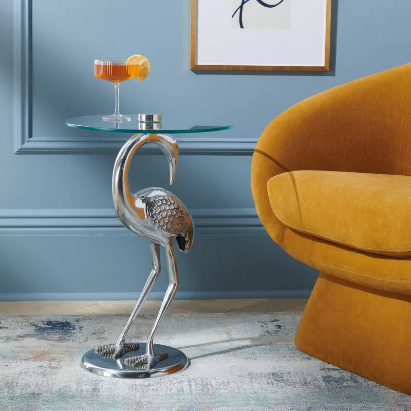 Silver flamingo-shaped side table with a cocktail in a room with a blue wall and yellow chair.