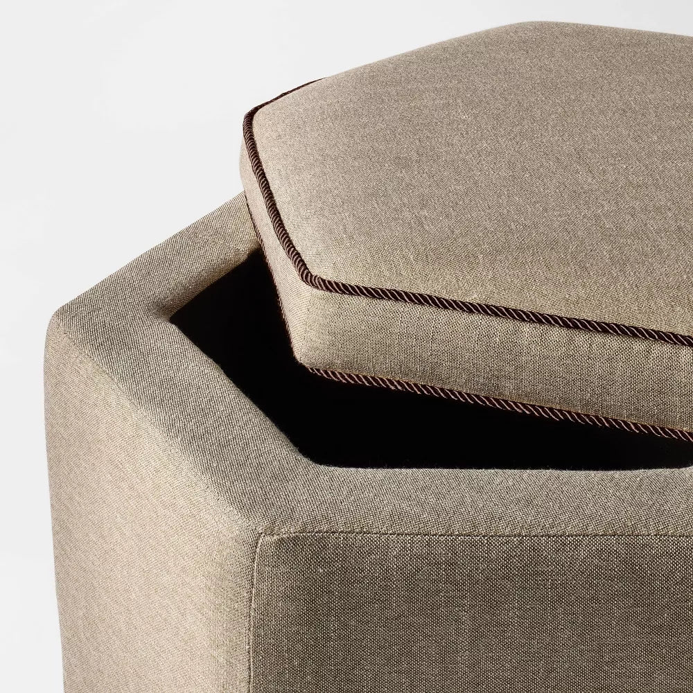 Hexagonal light gray colored ottoman with dark trim placed on a patterned rug in a living room setting.