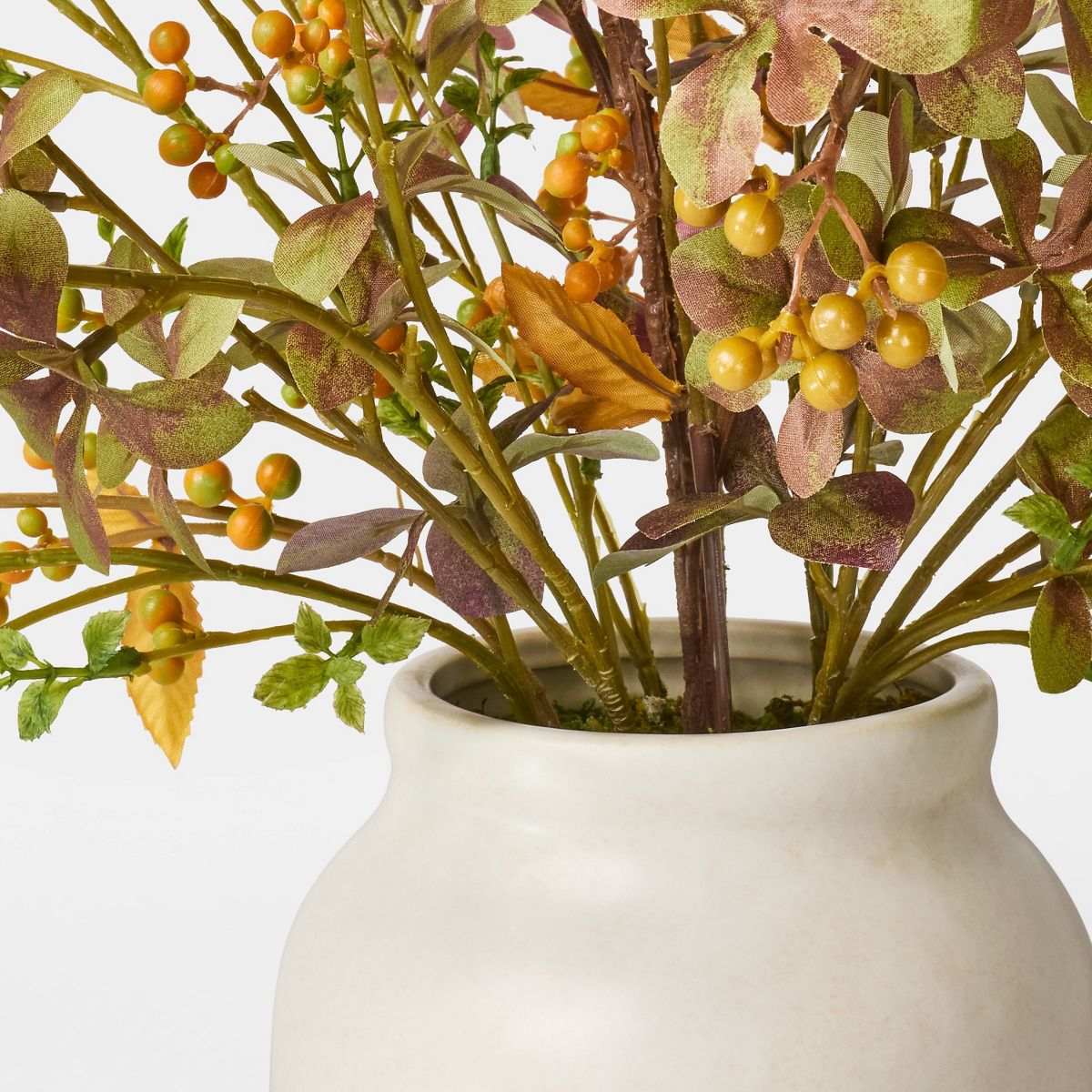 An artificial autumn leafy branch arrangement in a matte cream ceramic vase, photographed on a clean white studio background.