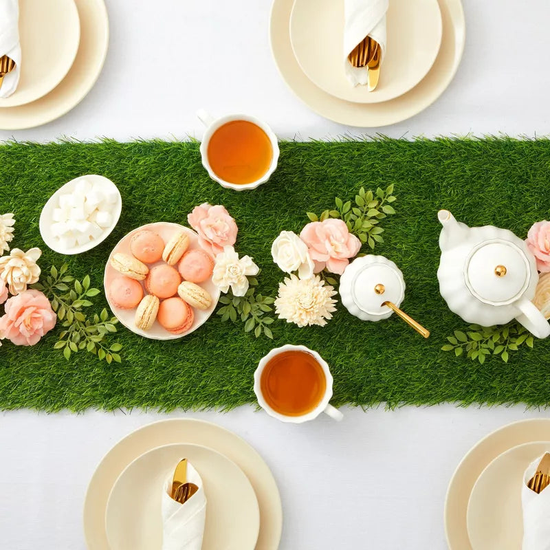 Tea party setup with green grass runner, teacups, cookies, and flowers on a white table.
