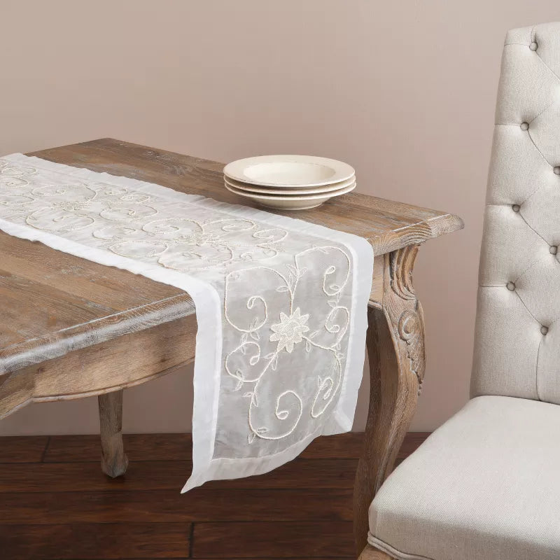 Wooden table with a white embroidered table runner and plates, next to a gray upholstered chair.
