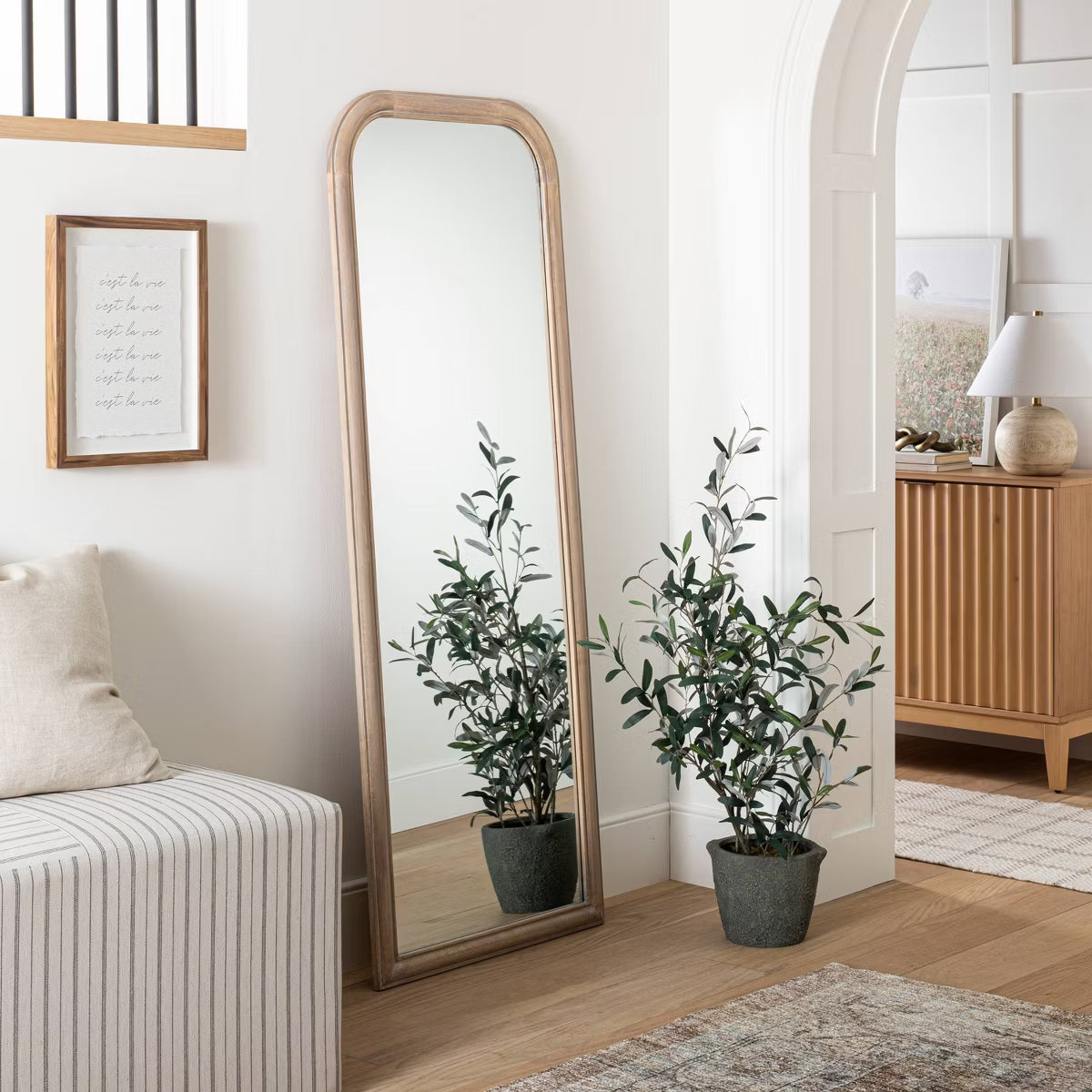 A full-length mirror with a gold colored frame is placed on the floor next to a wicker basket, a white potted plant, and a small basket with books.