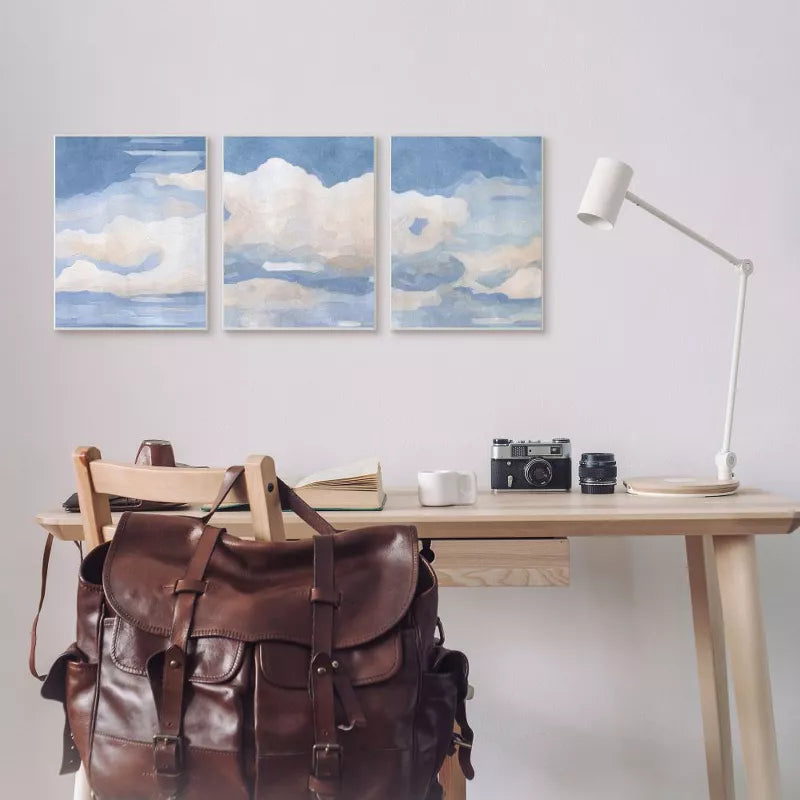 Desk with a brown leather backpack, camera, and lamp against a wall with three cloud paintings.