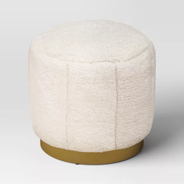 A round, cream colored ottoman with a textured surface and a gold colored base."