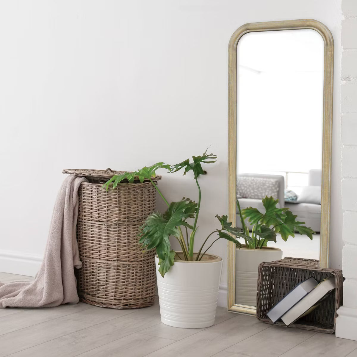 A full-length mirror with a gold colored frame is placed on the floor next to a wicker basket, a white potted plant, and a small basket with books.