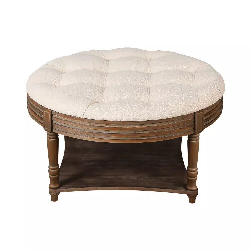 Round wooden coffee table with a beige cushion, vase of green flowers, and books on a patterned rug. beige