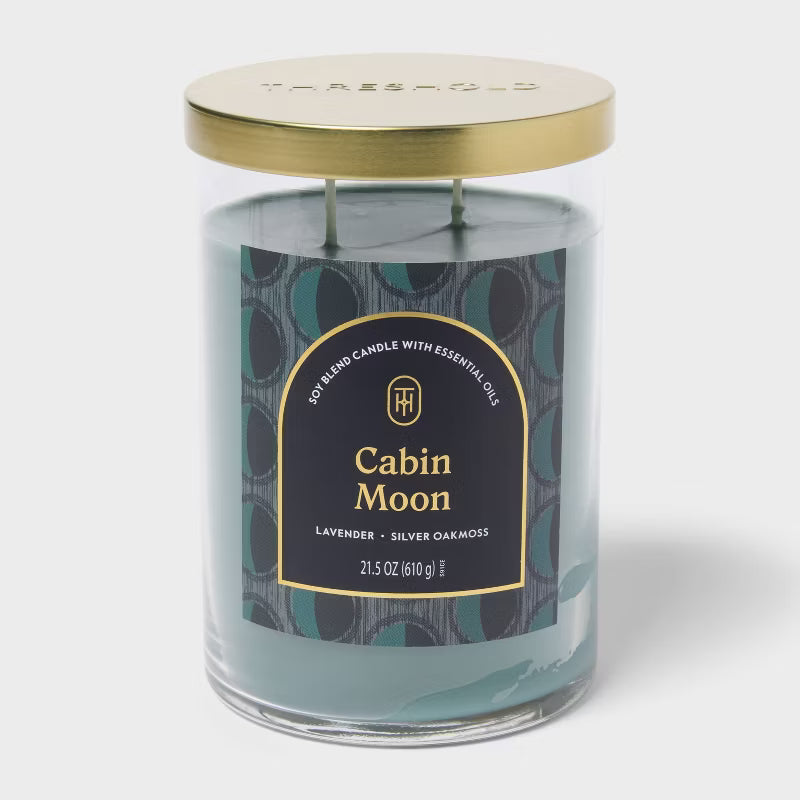 Candle labeled 'Cabin Moon' on a table with a vase and decorative items in the background 21.5oz