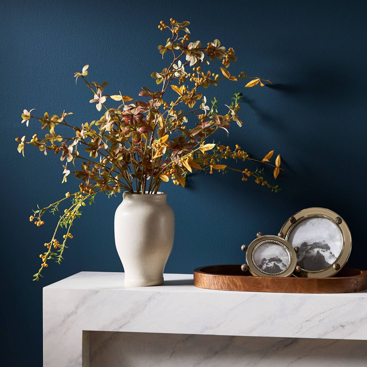 An artificial autumn leafy branch arrangement in a matte cream ceramic vase, photographed on a clean white studio background.