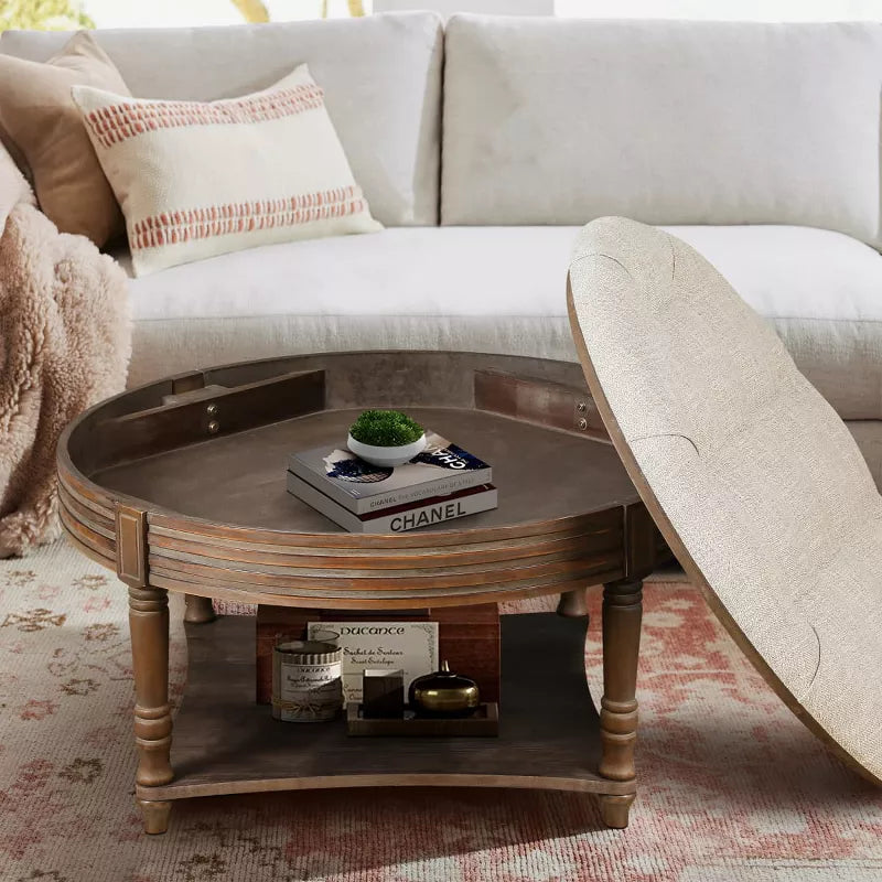 Round wooden coffee table with a beige cushion, vase of green flowers, and books on a patterned rug. beige