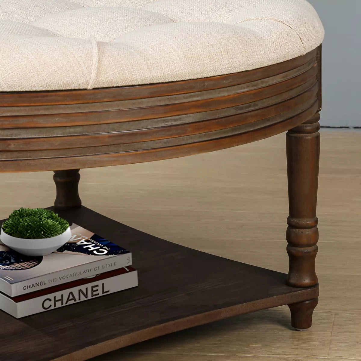 Round wooden coffee table with a beige cushion, vase of green flowers, and books on a patterned rug. beige