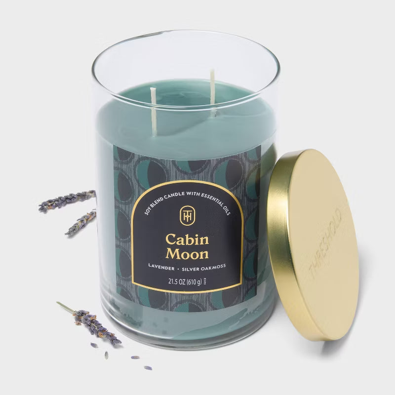 Candle labeled 'Cabin Moon' on a table with a vase and decorative items in the background 21.5oz