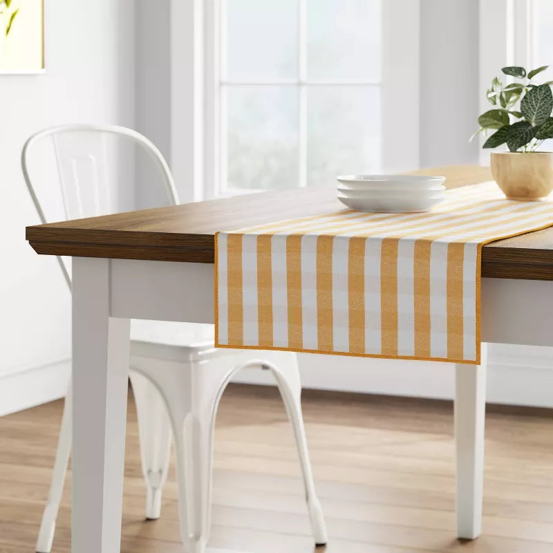 Dining table with a yellow and white striped runner, white chairs, and a plant in a bright room.
