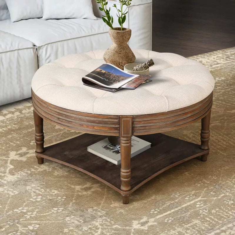 Round wooden coffee table with a beige cushion, vase of green flowers, and books on a patterned rug. beige