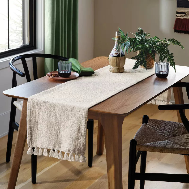 Dining table with a beige runner, plants, and glasses in a room with a window and curtains.