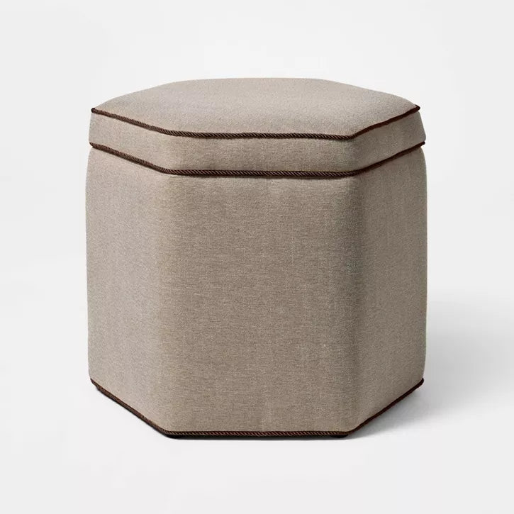 Hexagonal light gray colored ottoman with dark trim placed on a patterned rug in a living room setting.