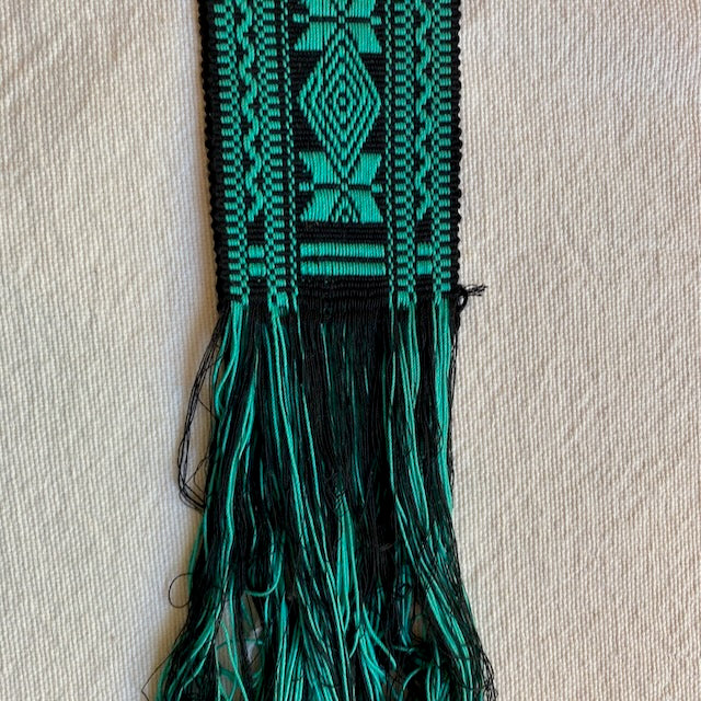 Green and black woven belt with tassels on a beige background Teal and black woven belt on a light gray fabric background Style G