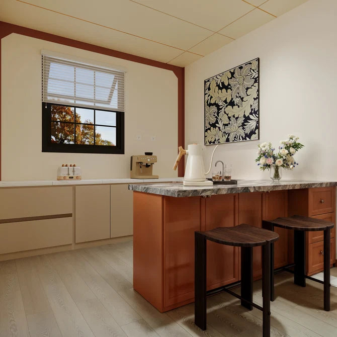 A modern kitchen interior with a central island in warm reddish-brown wood, a marble countertop, two dark wooden stools, cream walls, a window with blinds, and floral wall art.