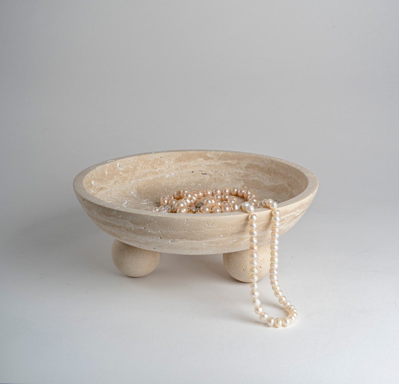 Beige ceramic bowl with three legs holding a pearl necklace on a light gray background