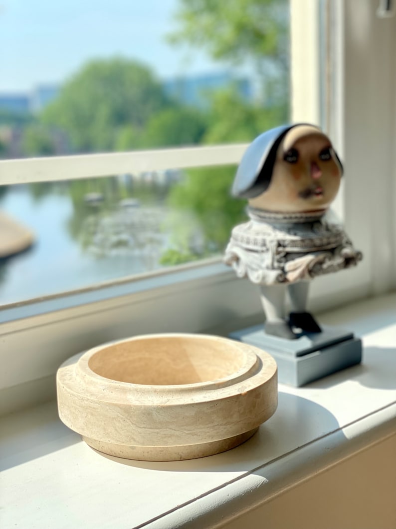 Wooden bowl and ceramic figure on a windowsill with a view of water and trees.