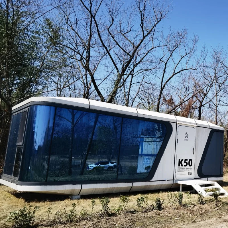 A modern white modular pod home with large dark blue-tinted windows sits outdoors on grassy ground among bare trees under a blue sky. The side of the unit shows the text K50 and a small white ramp at the front.