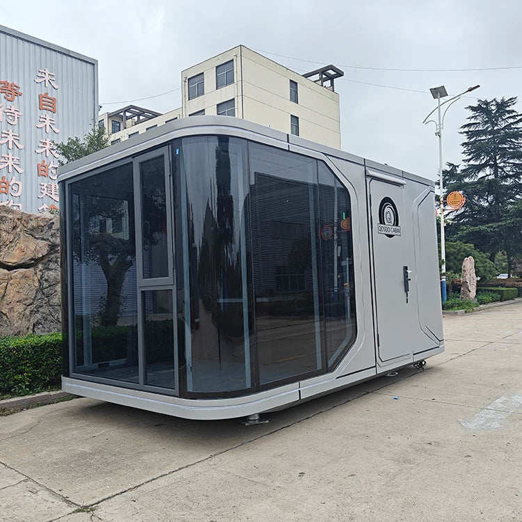 A modern modular mobile office or cabin with a silver-gray exterior and large dark-tinted glass windows, parked outdoors on a concrete surface near buildings and trees.