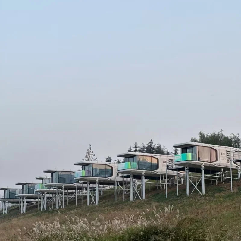 A row of modern elevated pods with large glass windows and white support structures, situated on a grassy hillside under a pale sky.