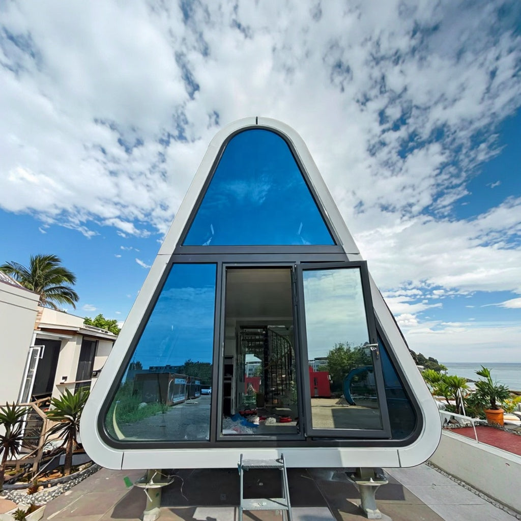 A modern A-frame glass cabin with a curved triangular roof and large reflective windows, shown outdoors against a blue sky with clouds. The structure has a white frame and is elevated on supports, with a small ladder in front.