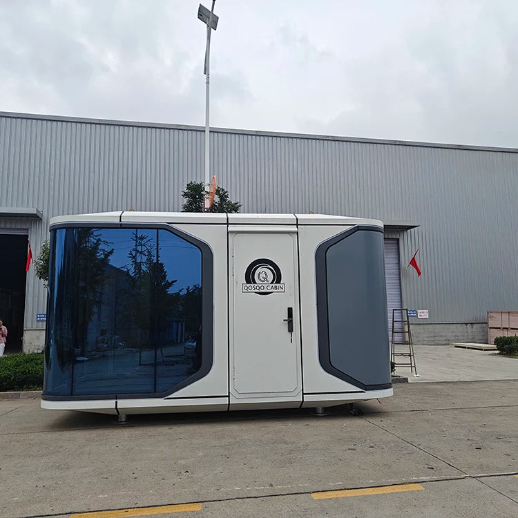 A small white and gray modular office pod with large tinted windows, parked outdoors in front of a gray metal warehouse building. The pod has a circular logo on the front door that appears to read 'DOC.