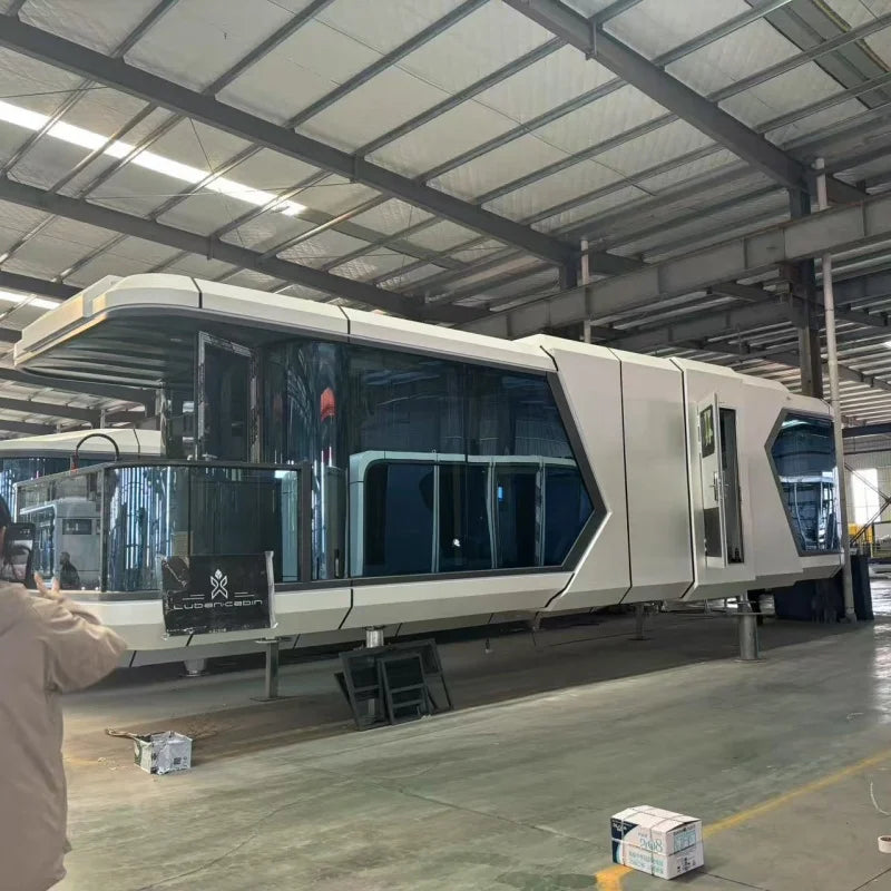 A large white modular pod or cabin with dark tinted windows sits indoors on a support frame inside a warehouse or factory. A person is partially visible on the left edge, and the industrial ceiling and floor are clearly visible.