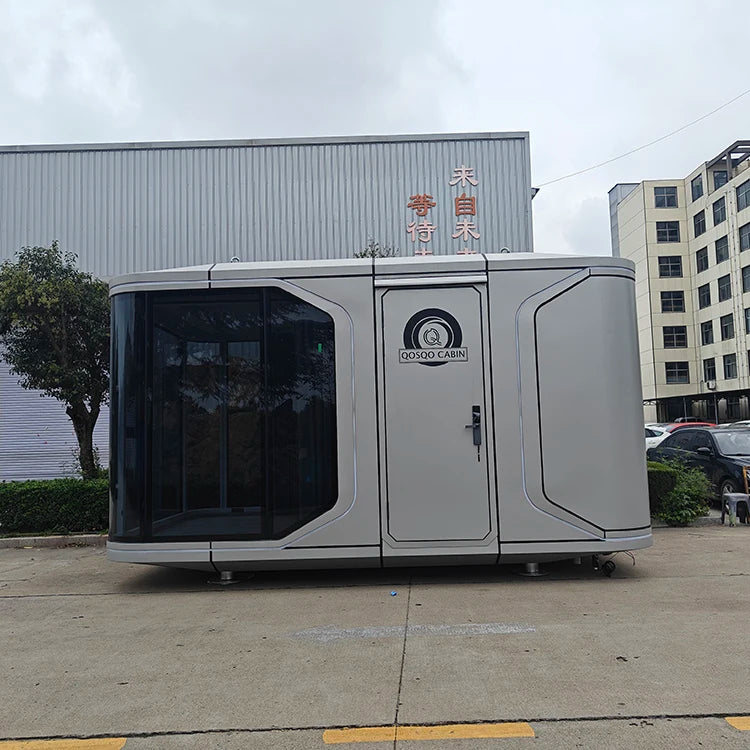 A compact, futuristic-looking gray container home with a large dark tinted glass window on the left and a central door labeled 'COSMO CARRY', parked outdoors in front of industrial buildings.