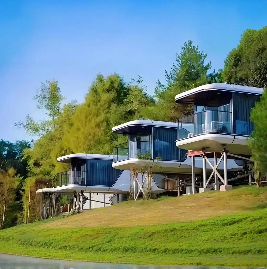 Modern floating treehouse-style cabins with large glass walls and elevated metal supports, set on a grassy hillside with dense green trees under a clear blue sky.