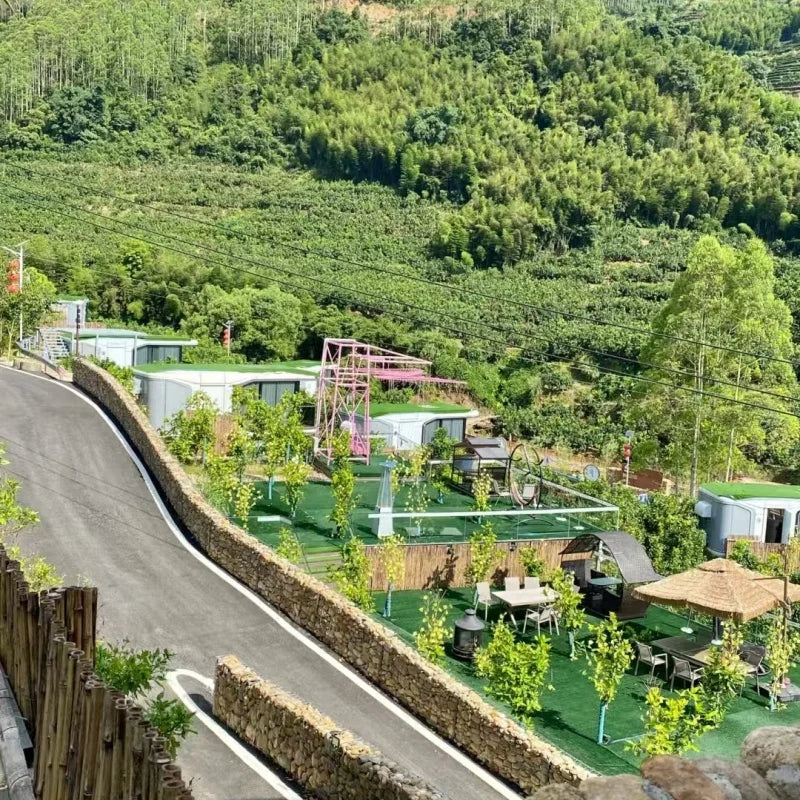 A scenic outdoor view of a mobile home park nestled on a lush green hillside. Several small modular homes with green roofs are arranged along a paved road, with patio seating, small trees, and a pink climbing structure nearby.