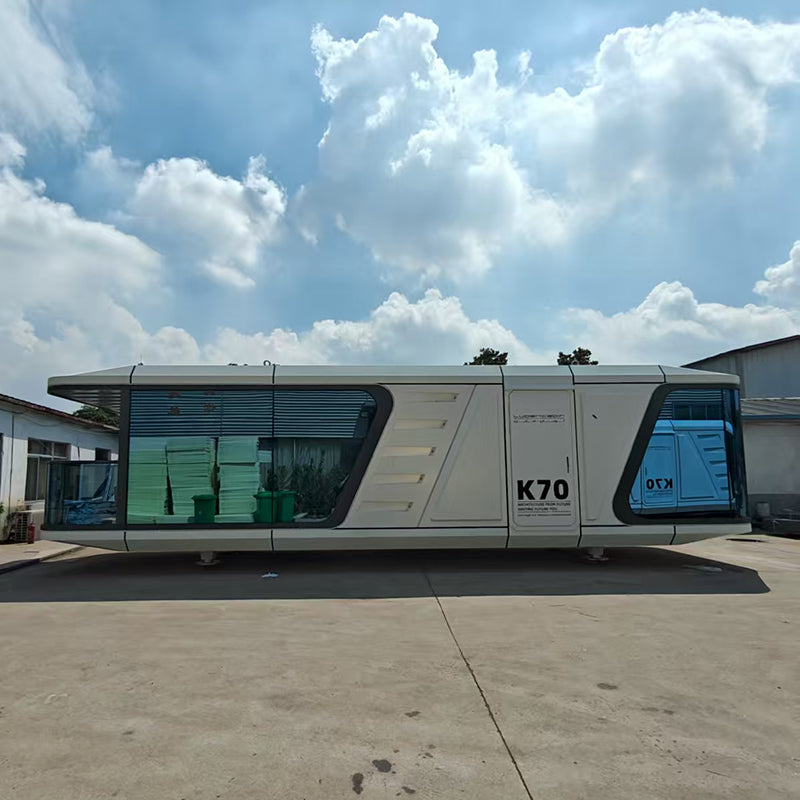 A modern white modular container home with large tinted windows, labeled K70, parked outdoors on a concrete surface under a blue sky with large white clouds.