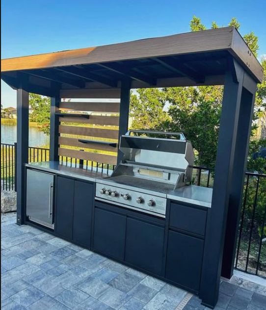Outdoor kitchen island with a built-in stainless steel gas grill under a dark wood and black pergola-style roof, set on a stone patio with a lake and trees in the background.
